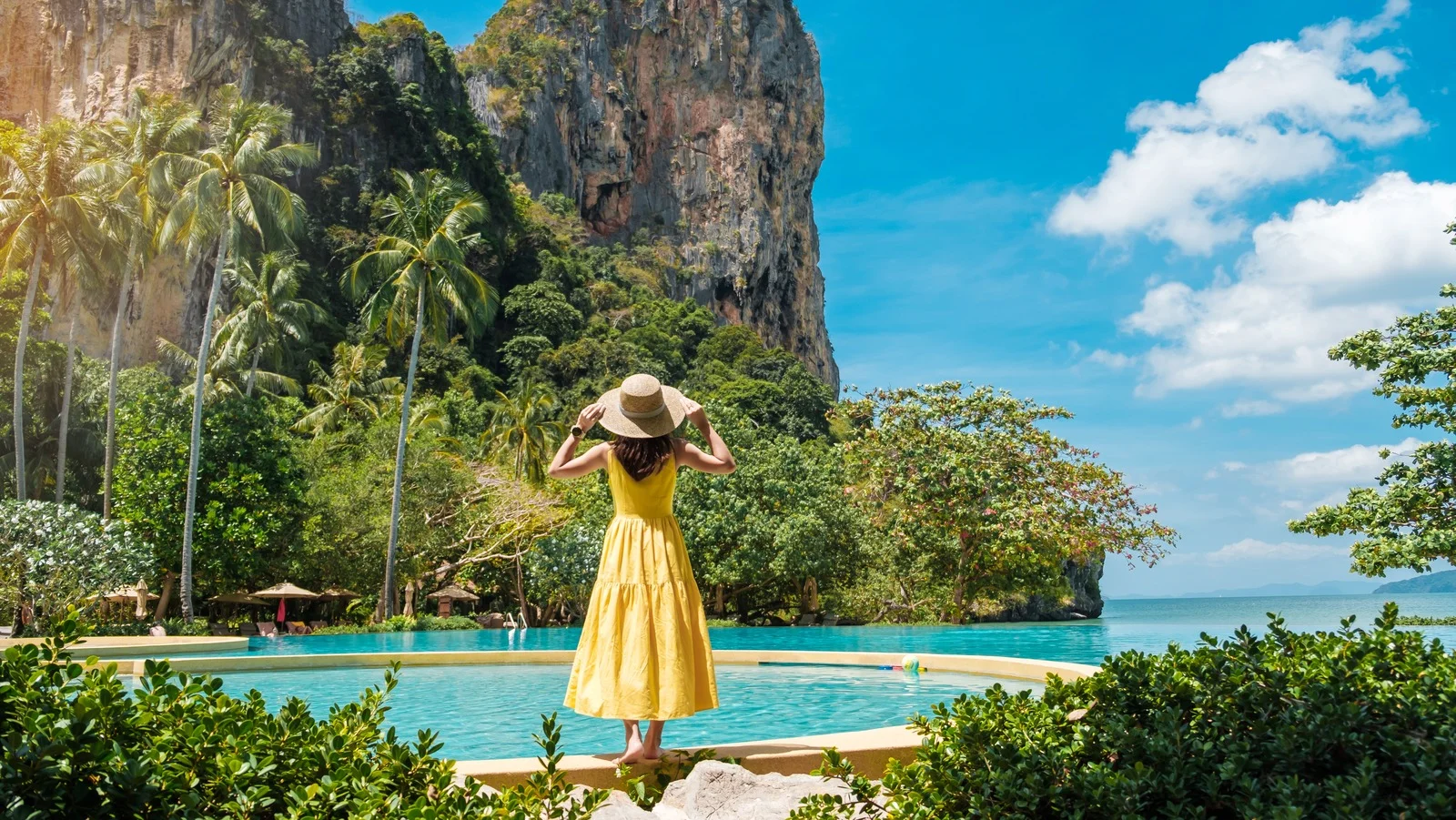 Ảnh bảo vệ môi trường với cô gái mặc váy vàng đứng bên hồ bơi giữa thiên nhiên nhiệt đới, biển xanh và vách đá hùng vĩ.